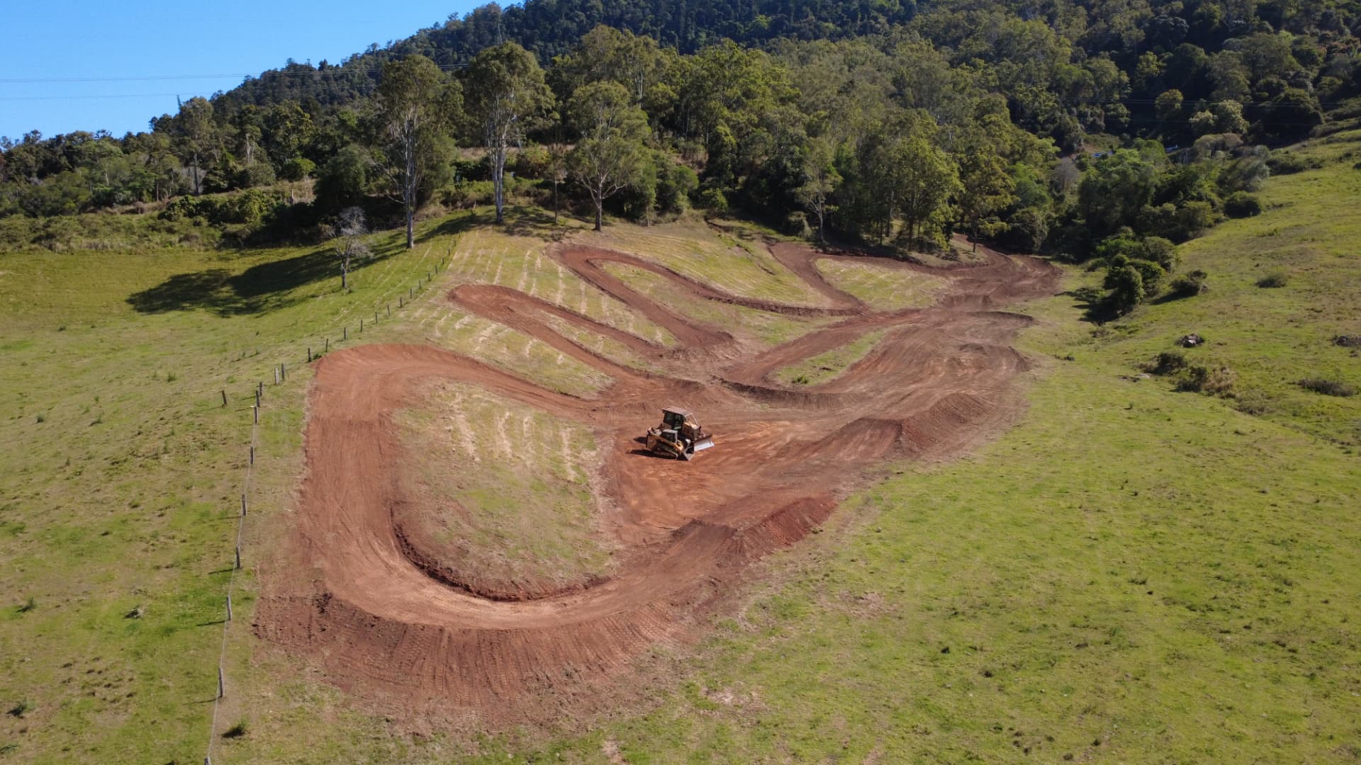 Aerial view of a partially built motocross track winding through green countryside with a bulldozer shaping the ground